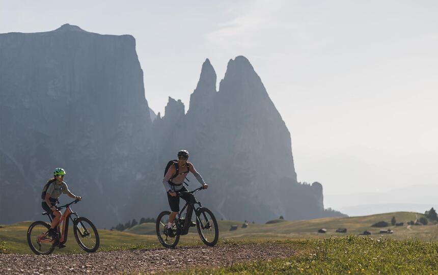 Zwei Mountainbiker fahren auf einem Fahrradweg; im Hintergrund erhebt sich der Schlern