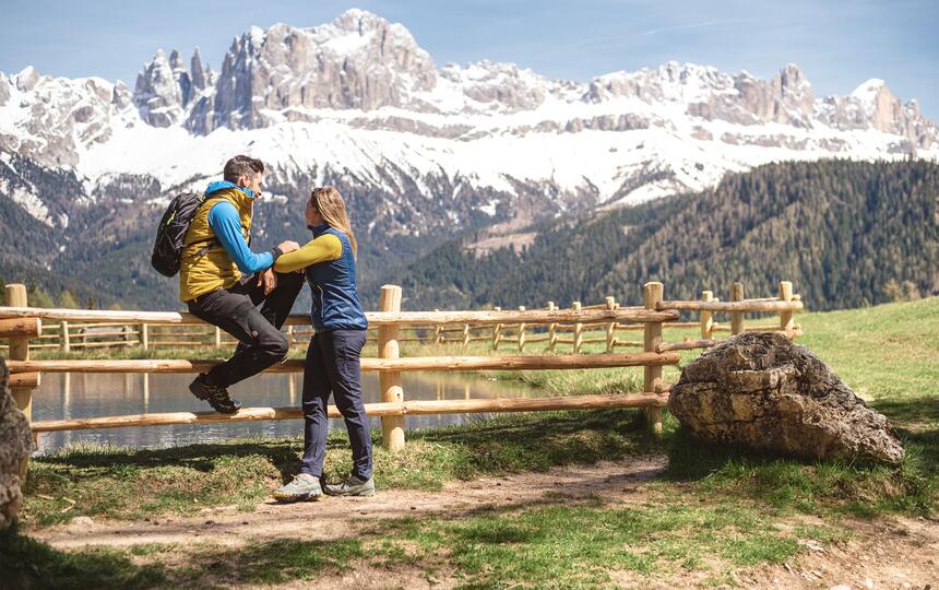 Ein Mann sitzt auf einem Zaun an einem See, eine Frau steht daneben, beide schauen auf die Dolomiten.