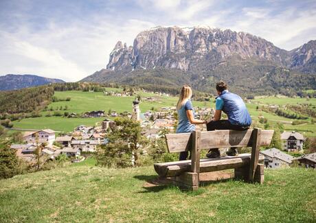 Ein Mann und eine Frau sitzen auf einer Holzbank und blicken auf Völs am Schlern