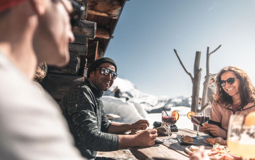 Mehrere Personen sitzen an einem Holztisch im Freien vor einer Berghütte im Schnee und trinken einen Aperitif