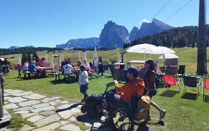 Mehrere Personen sitzen und stehen auf einer Wiese auf der Seiser Alm, ein Mann im Rollstuhl befindet sich im Vordergrund, im Hintergrund die Dolomiten
