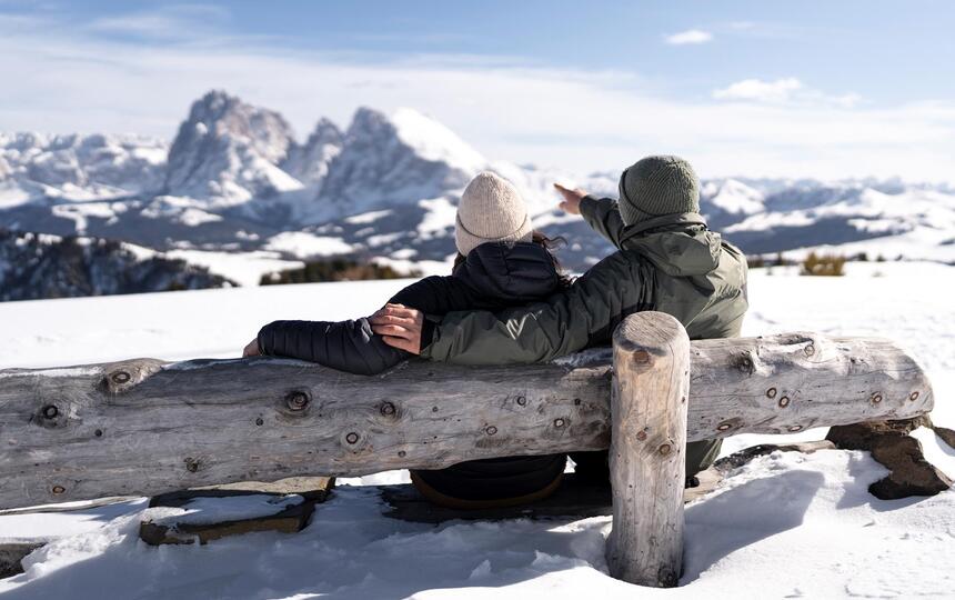 Ein Mann und eine Frau sitzt auf einer Holzbank und blickt auf die Dolomiten