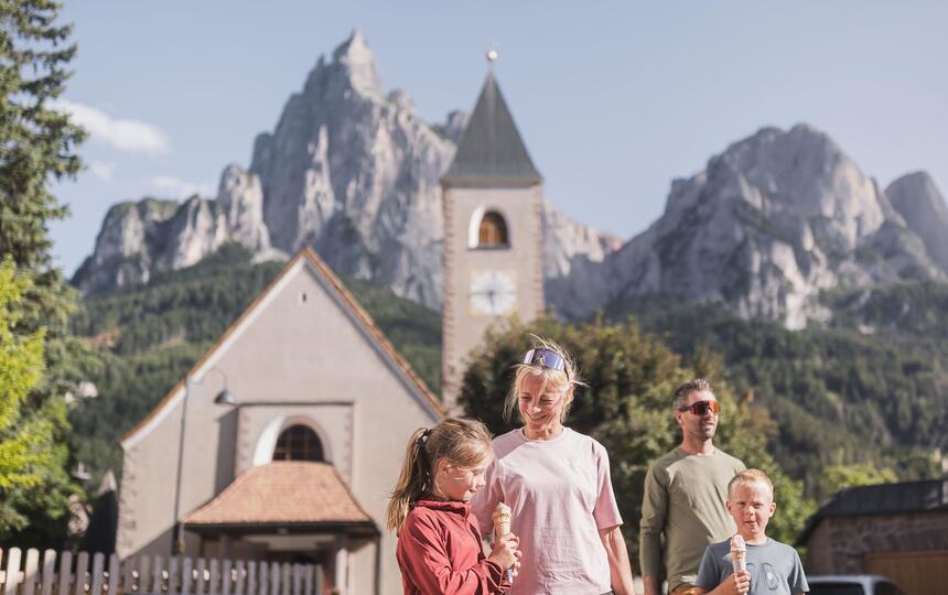 Una famiglia con due bambini mangia un gelato, con una chiesa e le Dolomiti sullo sfondo