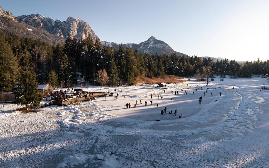 Luftbild des zugefrorenen Völser Weihers; zahlreiche Personen laufen darauf Schlittschuh