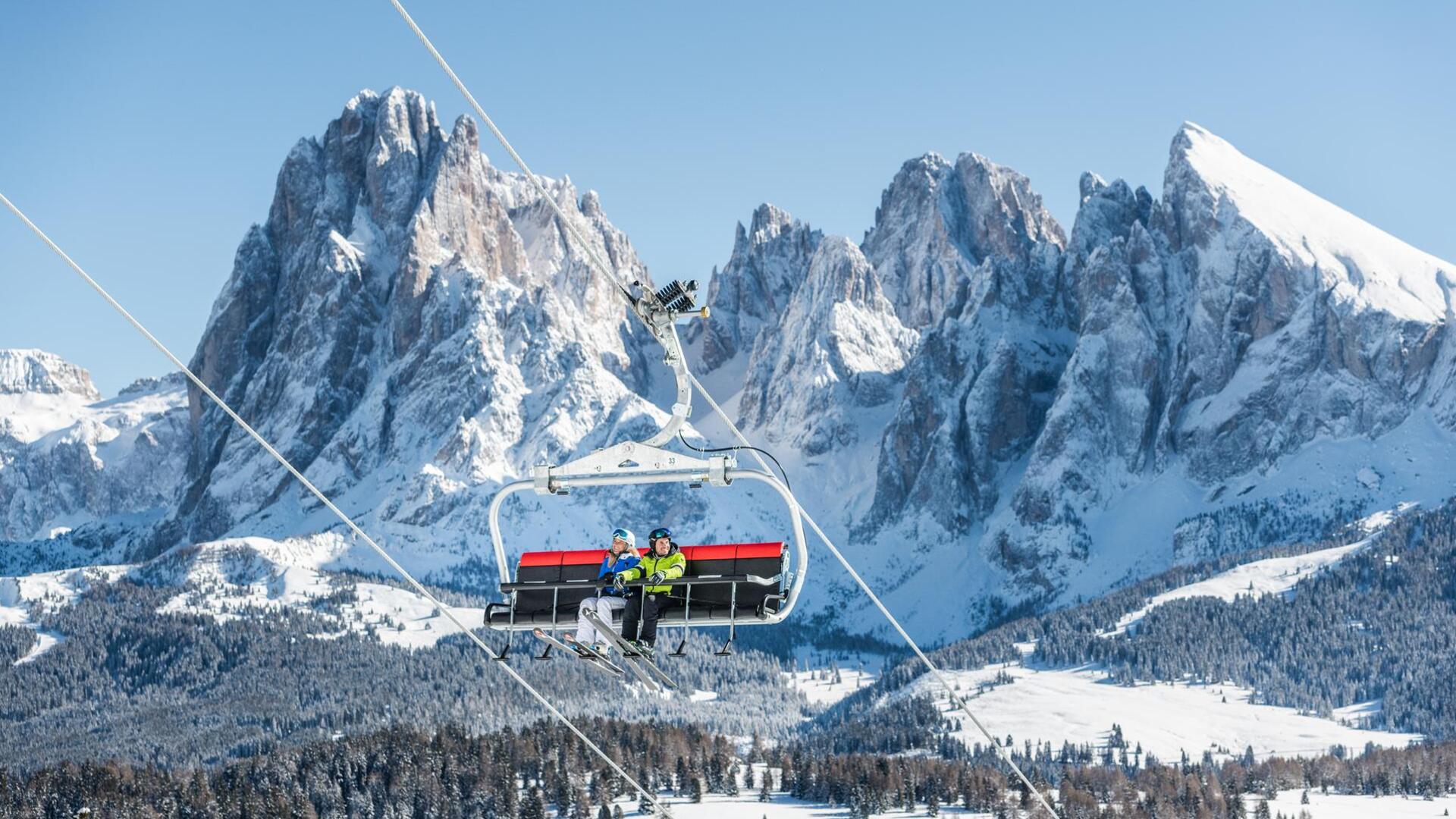 Ein Mann und eine Frau fahren mit dem Sessellift hoch, im Hintergrund der Plattkofel und der Langkofel