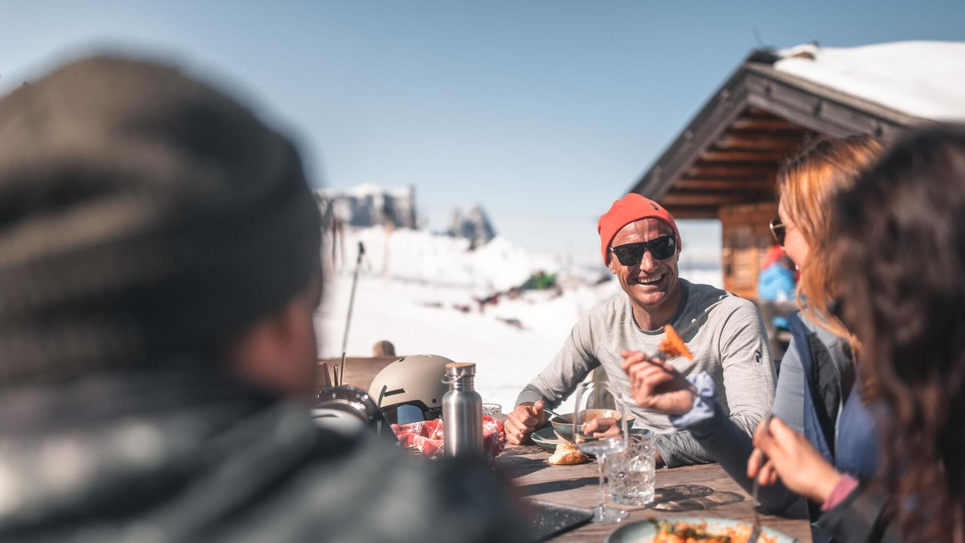 Eine Gruppe von Personen sitzt draußen an einem Holztisch und isst ein Nudelgericht.