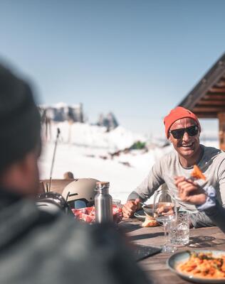 Eine Gruppe von Personen sitzt draußen an einem Holztisch und isst ein Nudelgericht.