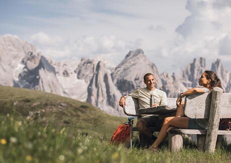Ein Mann und eine Frau sitzen auf einer Holzbank mit Blick auf die Dolomiten