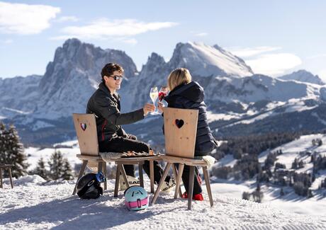 Two people sit outside, toasting with an aperitif, with snacks on the wooden table before them