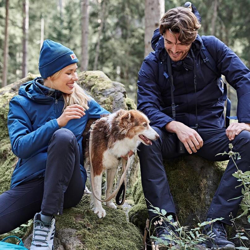 A man and a woman are sitting on a rock in the forest, next to them a dog is sitting