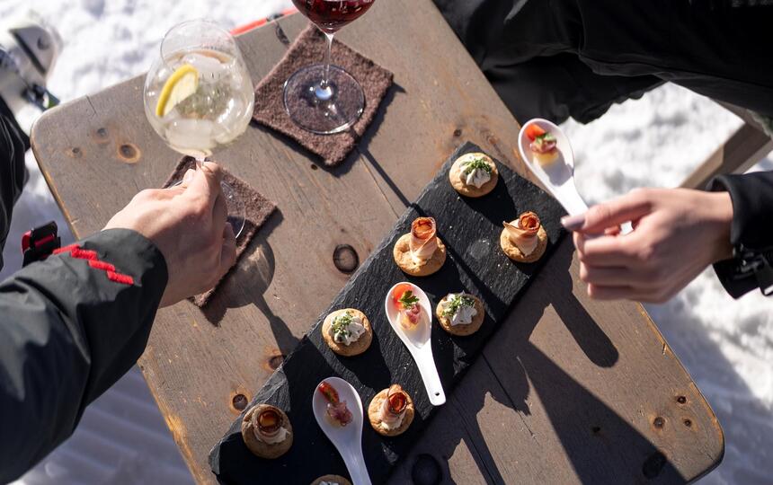 Mehrere Personen stehen im Schnee um einen kleinen Holztisch, greifen nach Snacks und stoßen mit Getränken an