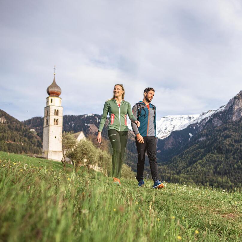 Ein Mann und eine Frau spazieren einer Wiese entlang, im Hintergrund eine Kirche