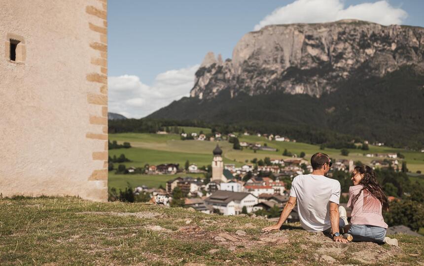 Ein Mann und eine Frau sitzen auf einer Wiese und blicken auf Völs am Schlern